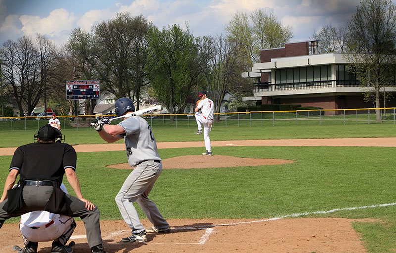 A photo of a MacMurray pitcher facing the camera throwing to a visiting team's batter in the foreground.